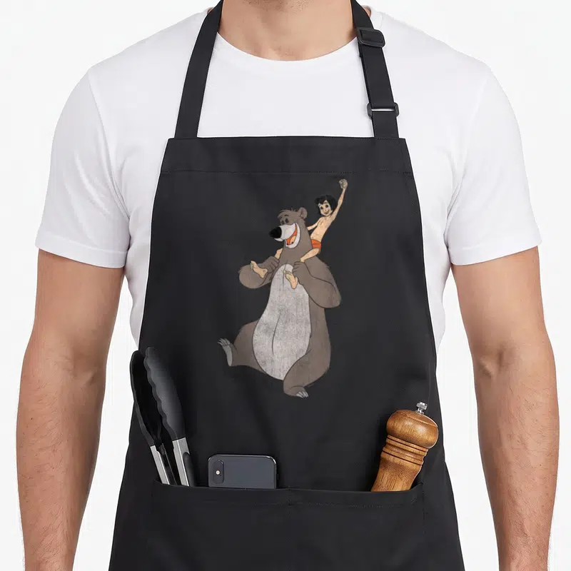Tablier de cuisine noir avec une illustration colorée de Mowgli sur les épaules de Baloo et leur signature en bas.