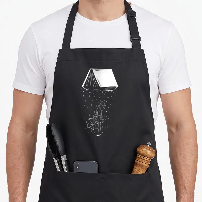 Tablier de cuisine noir avec une illustration blanche d'une fille tenant un parapluie sous un livre qui déverse des lettres comme de la pluie.