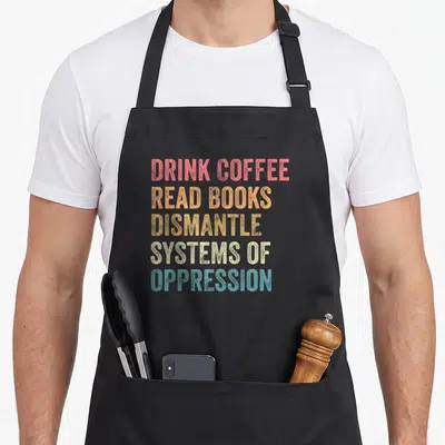 Tablier de cuisine noir avec texte multicolore Drink coffee read books dismantle systems of oppression imprimé au centre.