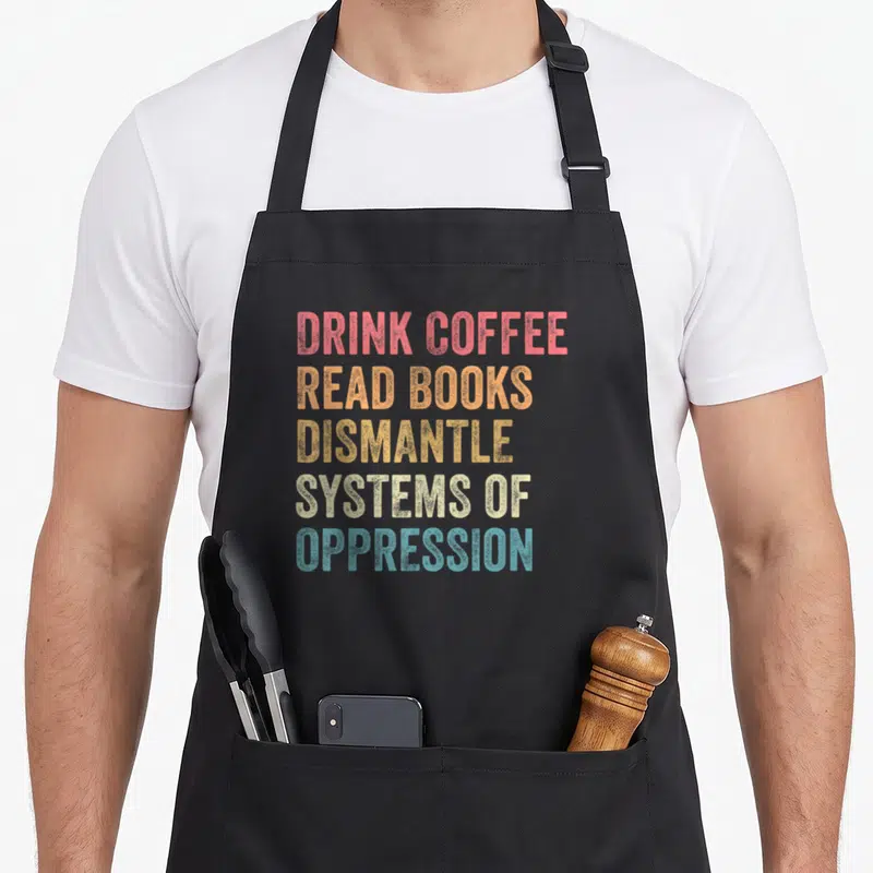 Tablier de cuisine noir avec texte multicolore Drink coffee read books dismantle systems of oppression imprimé au centre.