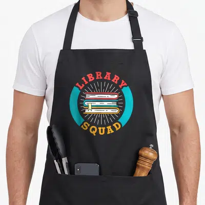 Tablier de cuisine noir avec logo Library Squad illustrant trois livres empilés sous des rayons lumineux.