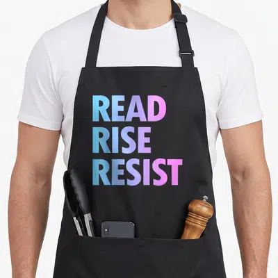 Tablier de cuisine noir avec le texte Read Rise Resist en lettres capitales aux couleurs dégradées bleu et rose.