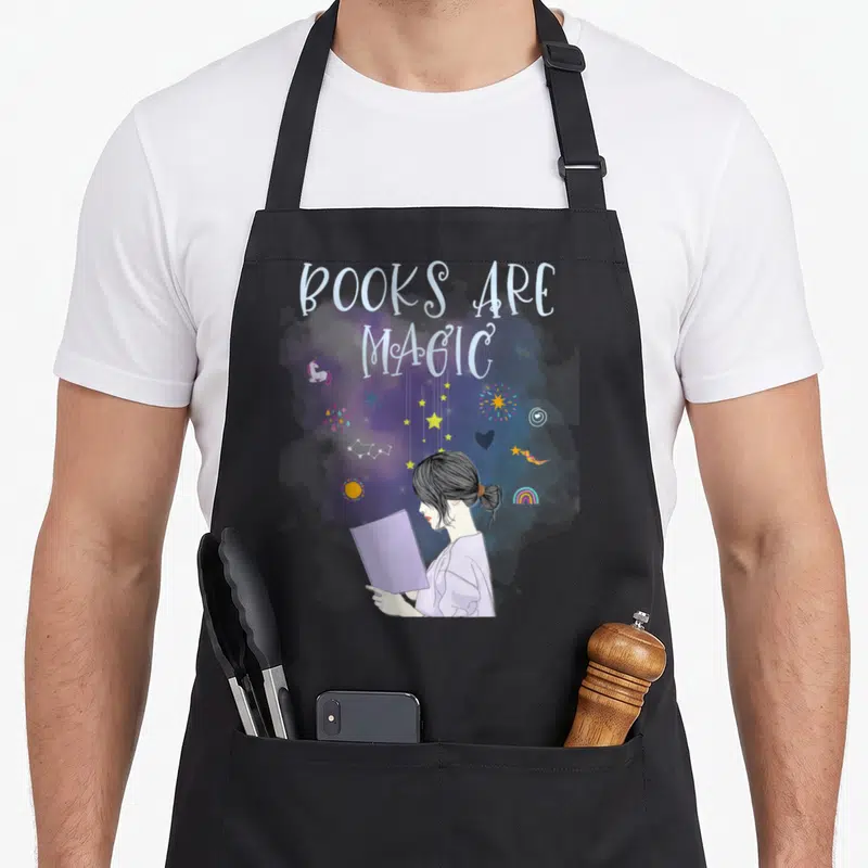 Tablier de cuisine noir avec une illustration d'une fille lisant un livre émettant des étoiles et des symboles magiques sous le texte Books Are Magic.