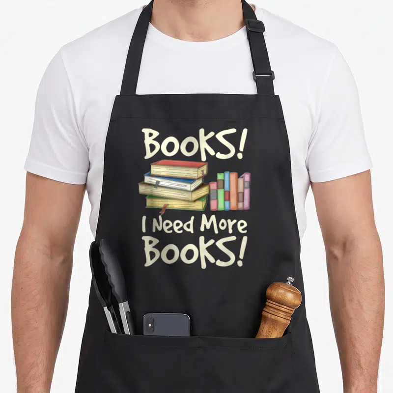 Tablier de cuisine noir avec illustration de livres empilés et texte Books I Need More Books en lettres crème.
