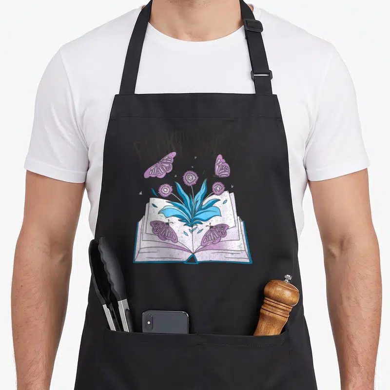 Tablier de cuisine noir avec un livre ouvert émergeant de fleurs bleues et de papillons violets sous le mot Booktrovert.