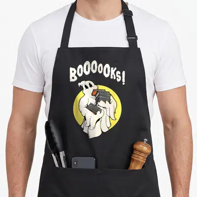 Tablier de cuisine noir avec un fantôme blanc tenant des livres devant un cercle jaune et le texte BOOOOOKS en haut.