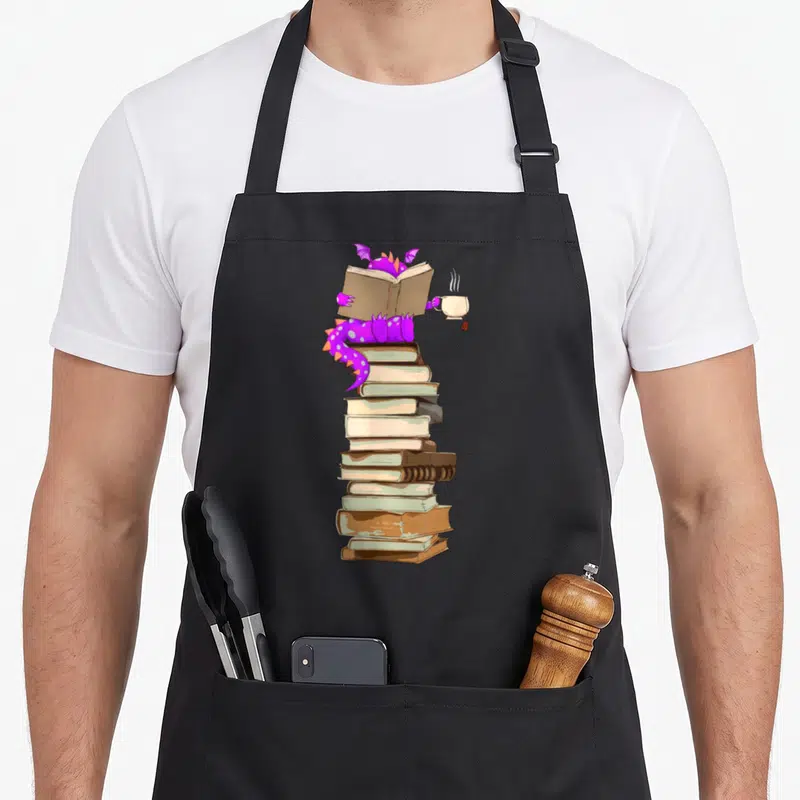 Tablier de cuisine noir avec un dragon violet lisant un livre sur une pile de volumes tout en tenant une tasse de thé.