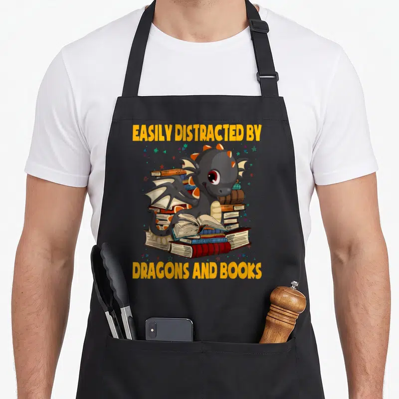 Tablier de cuisine noir avec un dragon gris lisant sur des livres colorés et des étoiles.