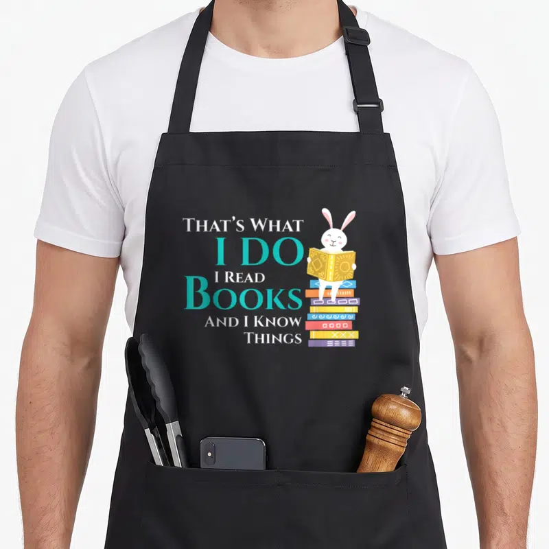 Tablier de cuisine noir avec un lapin blanc lisant sur une pile de livres multicolores et du texte typographié.