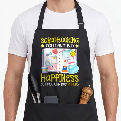 Tablier de cuisine noir avec illustration de scrapbook, ciseaux, colle et texte jaune sur fond gris uni.