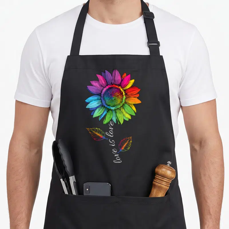Tablier de cuisine noir avec un grand tournesol aux couleurs de larc-en-ciel et deux plumes stylisées sous une inscription blanche.
