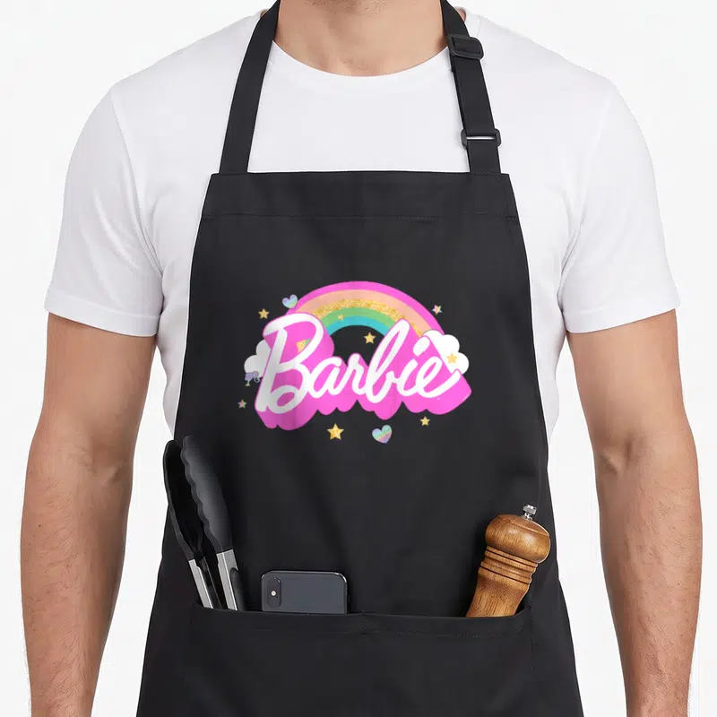 Tablier de cuisine noir avec le logo Barbie arc-en-ciel, des étoiles dorées et des cœurs colorés imprimés au centre.