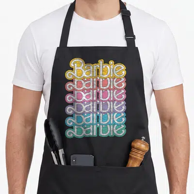 Tablier de cuisine noir avec le mot Barbie répété verticalement dans des teintes jaune, rose, violet et bleu pastel à effet texturé.