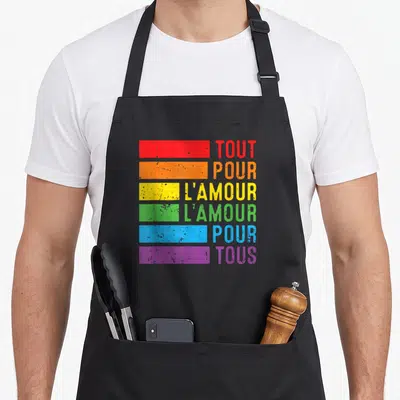 Tablier de cuisine noir avec bandes colorées arc-en-ciel et texte Tout Pour L'Amour pour Tous imprimé en gras.