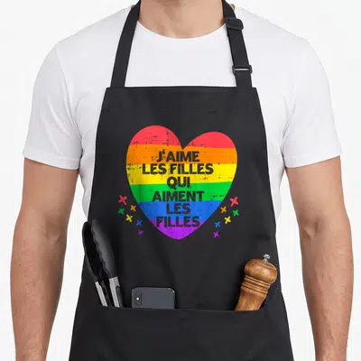 Tablier de cuisine noir avec un grand cœur arc-en-ciel portant l'inscription J'aime les filles qui aiment les filles entouré de petites étoiles colorées.