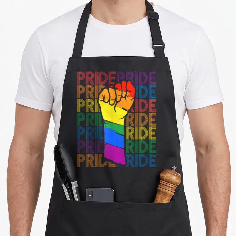 Tablier de cuisine noir avec un poing levé aux couleurs de la Pride et le mot Pride répété en arrière-plan typographique.