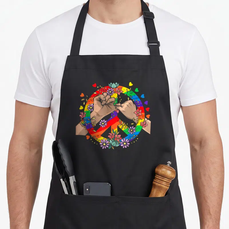 Tablier de cuisine noir avec illustration de poings jointifs dans un signe de paix arc-en-ciel décoré de fleurs et de cœurs.