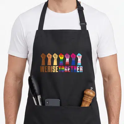 Tablier de cuisine noir avec sept poings levés multicolores et le texte We Rise Together Black Pride Equality imprimé en bas.