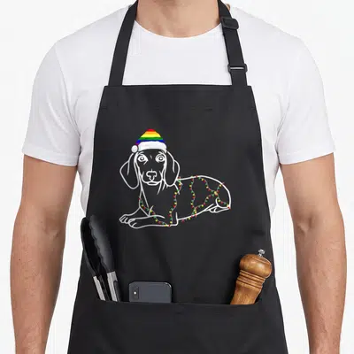 Tablier de cuisine noir avec illustration d'un teckel blanc portant une casquette arc-en-ciel et des lumières de Noël colorées.