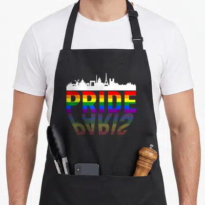 Tablier de cuisine noir avec l'inscription Pride en couleurs arc-en-ciel et un skyline blanc de Paris au-dessus.