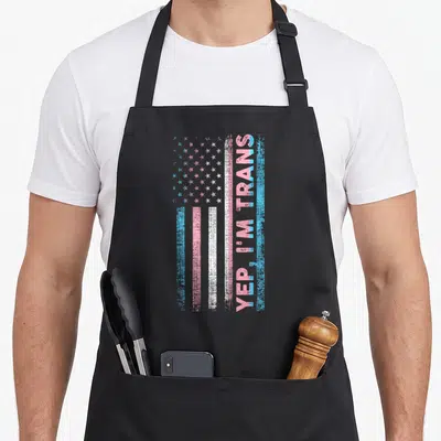 Tablier de cuisine noir avec un drapeau aux couleurs trans et le texte YEP I M TRANS imprimé verticalement sur le côté droit.