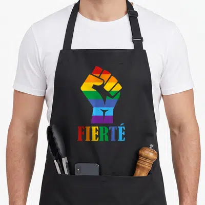 Tablier de cuisine noir avec un poing arc-en-ciel et le mot Fierté imprimé en lettres colorées.