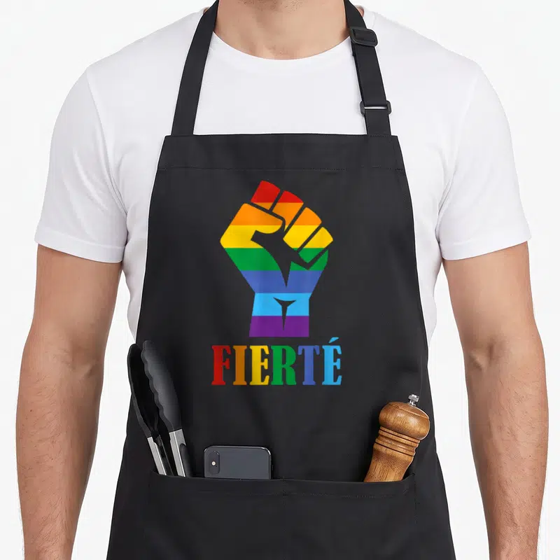 Tablier de cuisine noir avec un poing arc-en-ciel et le mot Fierté imprimé en lettres colorées.