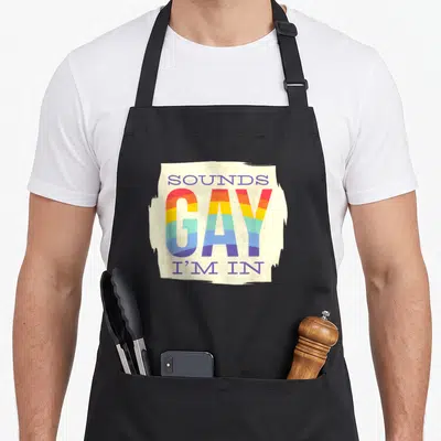 Tablier de cuisine noir avec un motif central Sounds Gay I'm In en lettres arc-en-ciel sur fond crème tacheté.