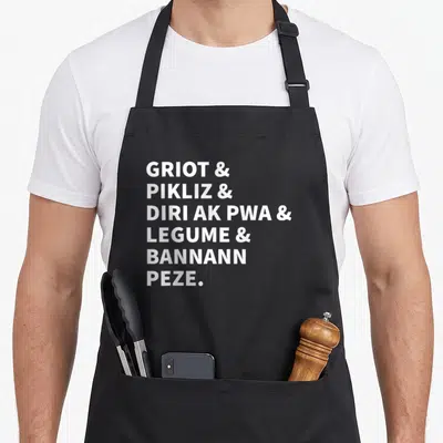Tablier de cuisine noir avec le texte blanc Griot et Pikliz et Dir ak Pwa et Legume et Bannann Peze centré sur le devant.