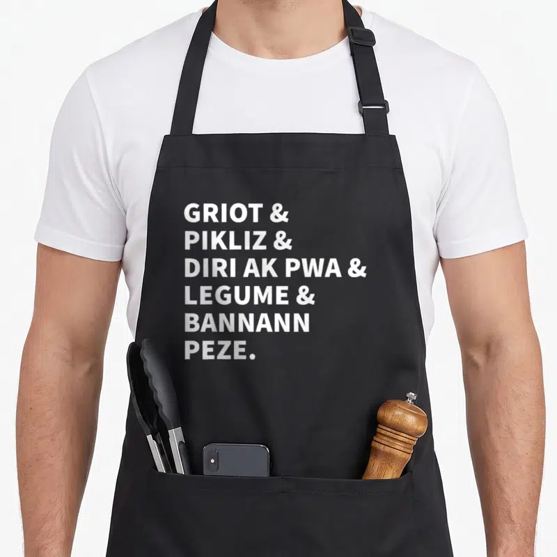 Tablier de cuisine noir avec le texte blanc Griot et Pikliz et Dir ak Pwa et Legume et Bannann Peze centré sur le devant.