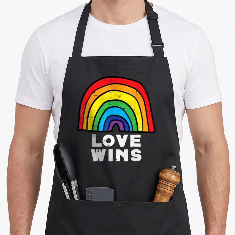 Tablier de cuisine noir avec arc-en-ciel coloré et texte blanc Love Wins en bas du motif central.
