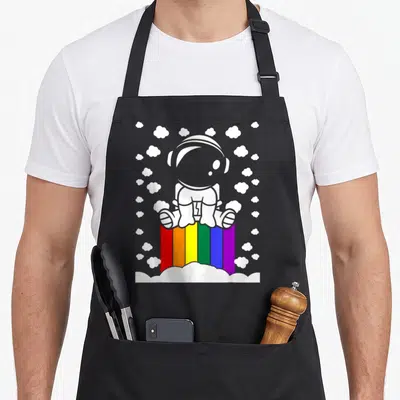 Tablier de cuisine noir avec illustration d'un astronaute blanc posé sur un arc-en-ciel multicolore parmi des nuages blancs.