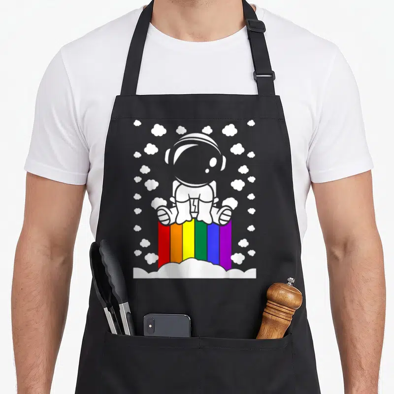 Tablier de cuisine noir avec illustration d'un astronaute blanc posé sur un arc-en-ciel multicolore parmi des nuages blancs.