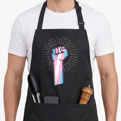 Tablier de cuisine noir avec un poing levé aux couleurs bleu, blanc et rose du drapeau transgenre.