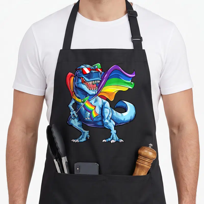 Tablier de cuisine noir avec illustration d'un dinosaure T rex bleu aux lunettes de soleil tenant un drapeau multicolore.