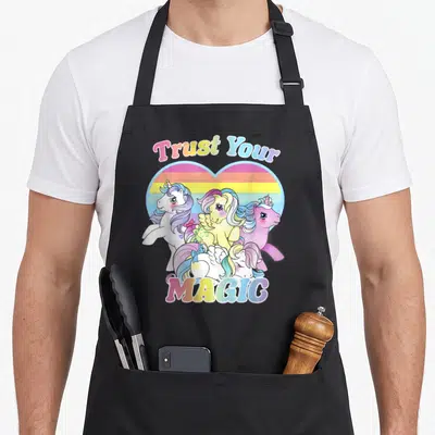 Tablier de cuisine noir avec des licornes colorées et le texte Trust Your Magic dans un cœur arc-en-ciel.
