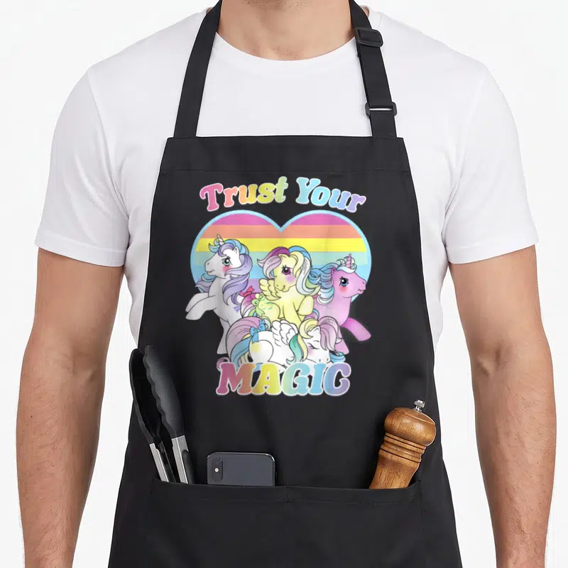 Tablier de cuisine noir avec des licornes colorées et le texte Trust Your Magic dans un cœur arc-en-ciel.