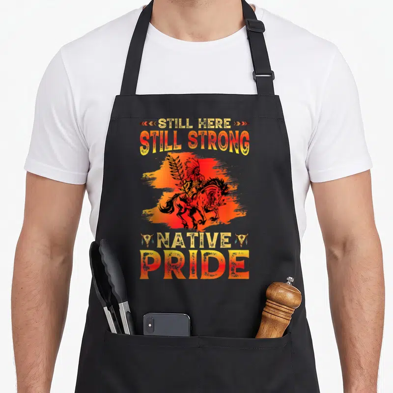 Tablier de cuisine noir avec illustration d'un guerrier amérindien à cheval et texte Still Strong Native Pride en dégradé orange.