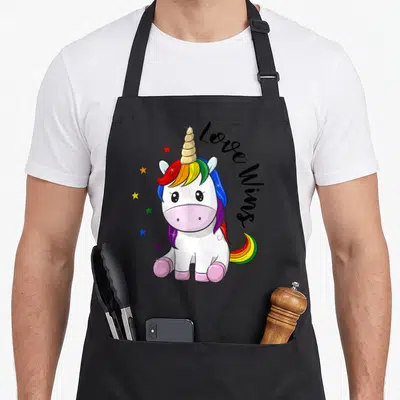Tablier de cuisine noir avec une licorne rose aux cheveux multicolores et les mots Love Wings écrits en noir à côté.