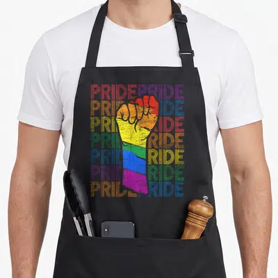 Tablier de cuisine noir avec un poing levé coloré et le mot Pride répété en arrière-plan typographique.