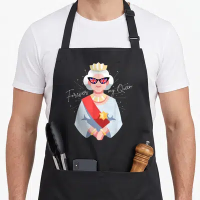 Tablier de cuisine noir avec illustration d'une reine aux verres Union Jack et texte cursive blanc.