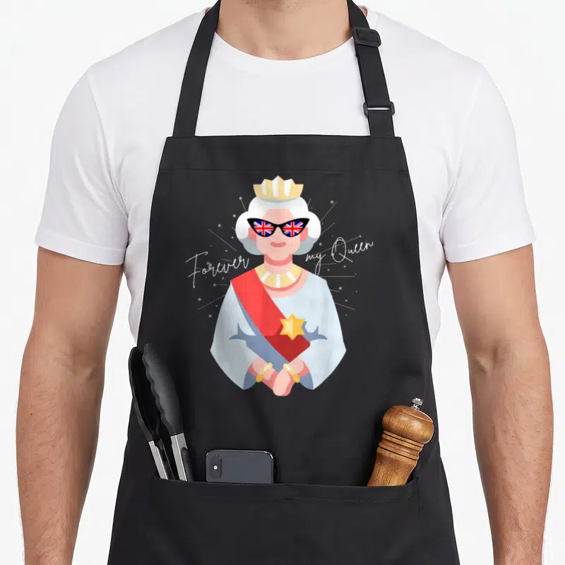 Tablier de cuisine noir avec illustration d'une reine aux verres Union Jack et texte cursive blanc.