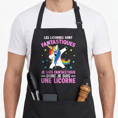 Tablier de cuisine noir avec licorne blanche aux cheveux arc-en-ciel effectuant un dab entourée d'étoiles colorées et de texte rose.