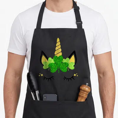 Tablier de cuisine noir avec un visage de licorne aux yeux fermés, une corne dorée et un trèfle vert pailleté au centre.