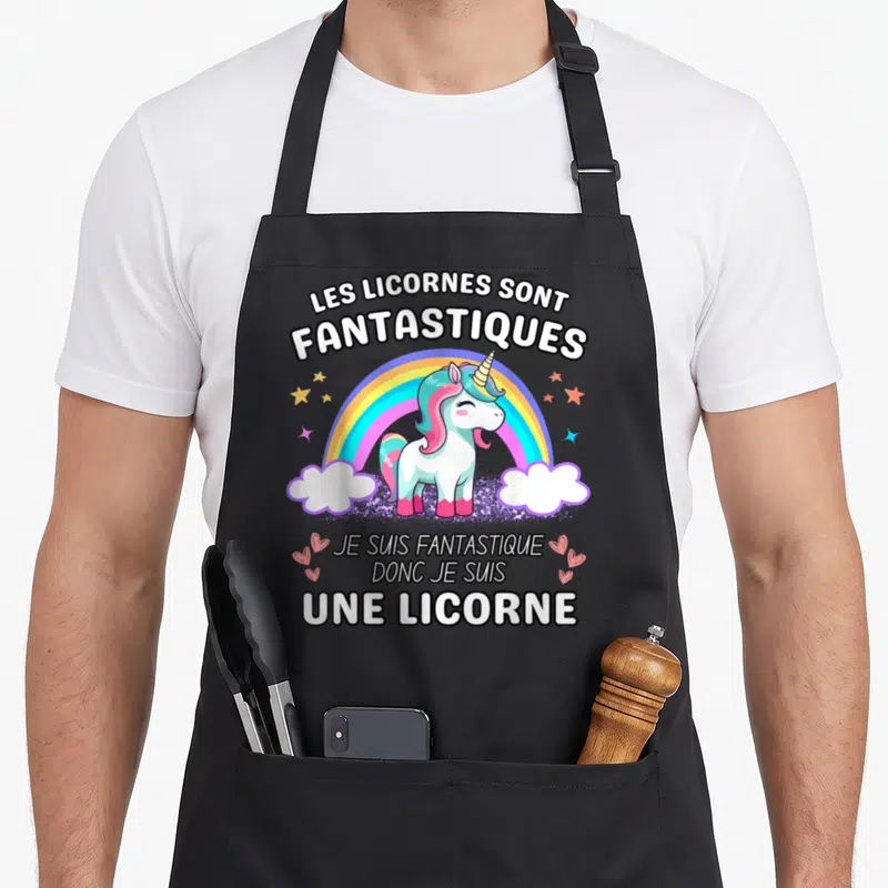 Tablier de cuisine noir avec illustration colorée d'une licorne blanche sous un arc-en-ciel et des étoiles dorées.