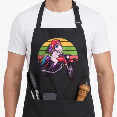 Tablier de cuisine noir avec illustration d'une licorne kawaii au casque rouge pilotant une moto rouge sur fond de soleil rétro aux bandes colorées.