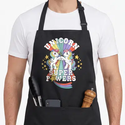 Tablier de cuisine noir avec deux licornes aux manés multicolores se touchant le front devant un arc-en-ciel rayonnant.