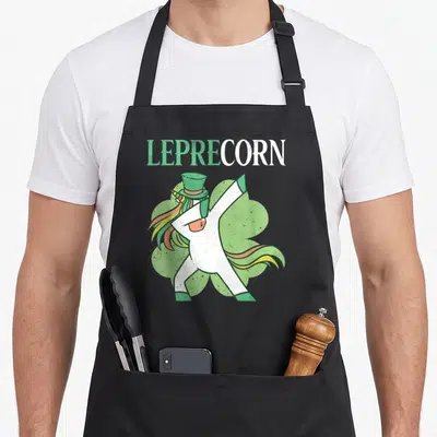Tablier de cuisine noir avec une licorne blanche en chapeau vert effectuant un mouvement de danse dab devant un trèfle.