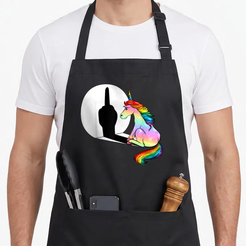 Tablier de cuisine noir avec licorne aux couleurs arc-en-ciel observant son ombre en geste sarcastique dans un miroir.