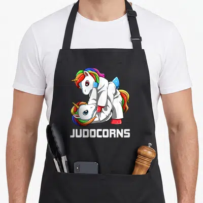 Tablier de cuisine noir avec deux licornes en kimono blanc pratiquant le judo sous le mot JUDOCORNS.